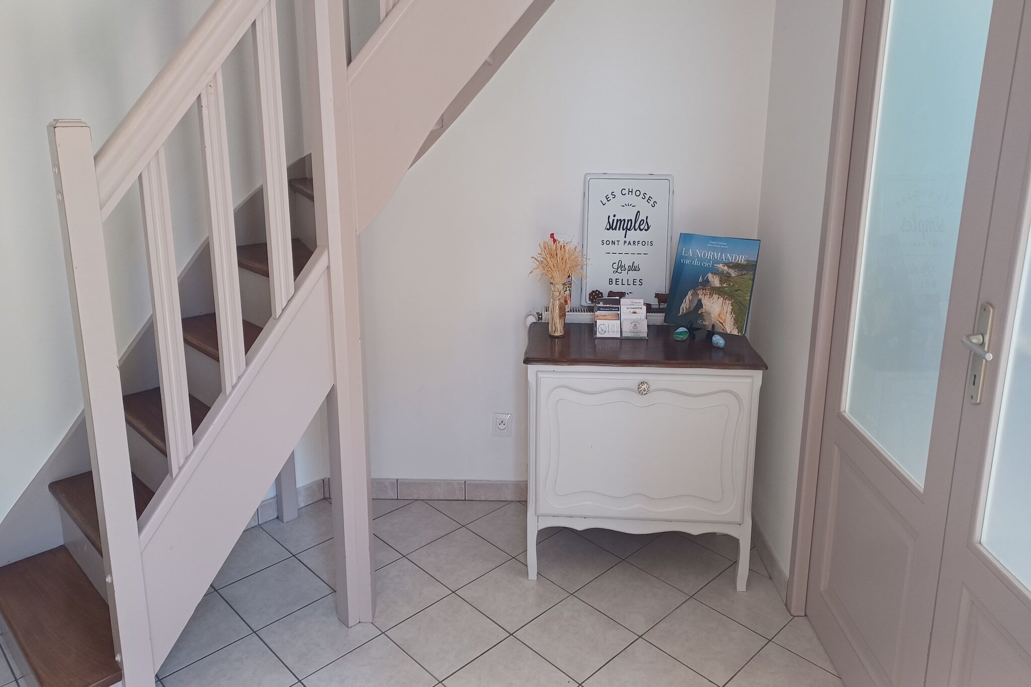 Un escalier avec des rampes blanches et des marches en bois mène à un palier. Sur ce palier, on trouve une console blanche ornée d’un vase décoratif et d’une enseigne.