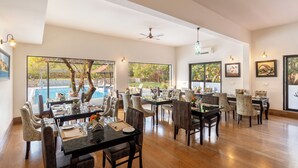 A spacious dining area with multiple tables and chairs, large windows offering views of a swimming pool and greenery, and ceiling fans. - The Hunting Lodge (Udaipur)