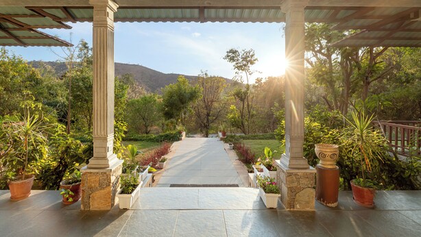 A covered pathway leading to a garden with potted plants and a view of hills. - The Hunting Lodge (Udaipur)