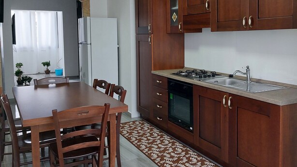 Cuina moderna amb armaris de fusta, fogó a gas i forn encastrat. Hi ha una taula de dinar amb cadires i una catifes amb dibuixos que condueix a la cuina.