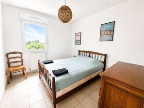 A bedroom with a bed, a chair, a bedside table, a hanging light fixture, and two framed pictures on the wall. - Spacious apartment - Balcony - 50m from the beach (Courseulles-sur-Mer)