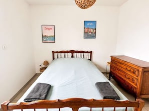 A bedroom with a wooden bed, a dresser, two framed pictures, and a lamp. - Spacious apartment – balcony – 50 m from the beach (Courseulles-sur-Mer)