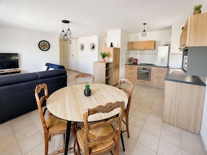 A modern kitchen with wooden cabinets, a dining table with chairs, and a living area with a sofa and television.