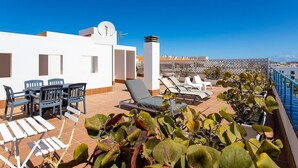 Eine Dachterrasse mit einem weißen Gebäude, einem Tisch mit Stühlen, Liegestühlen und Topfpflanzen.