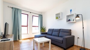 Ein Wohnzimmer mit grauem Sofa, einem weißen Couchtisch, einem Fernseher auf einem Holzständer und einer Stehlampe.