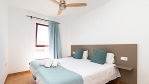 Un dormitorio con cama, ventilador de techo, ventana con cortinas y mesita de noche.