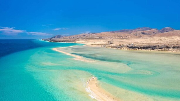 Un paisaje costero con aguas turquesas cristalinas, una playa de arena y montañas a lo lejos.
