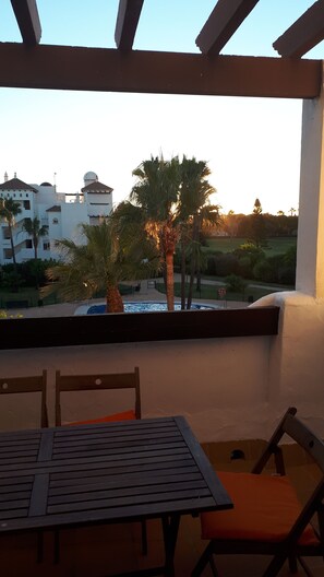 Un patio techado con una mesa y sillas, con vista a una piscina y palmeras, y edificios al fondo.