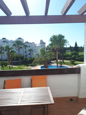 Un patio al aire libre con una mesa y sillas, con vista a una piscina y un área ajardinada con palmeras y un edificio al fondo.