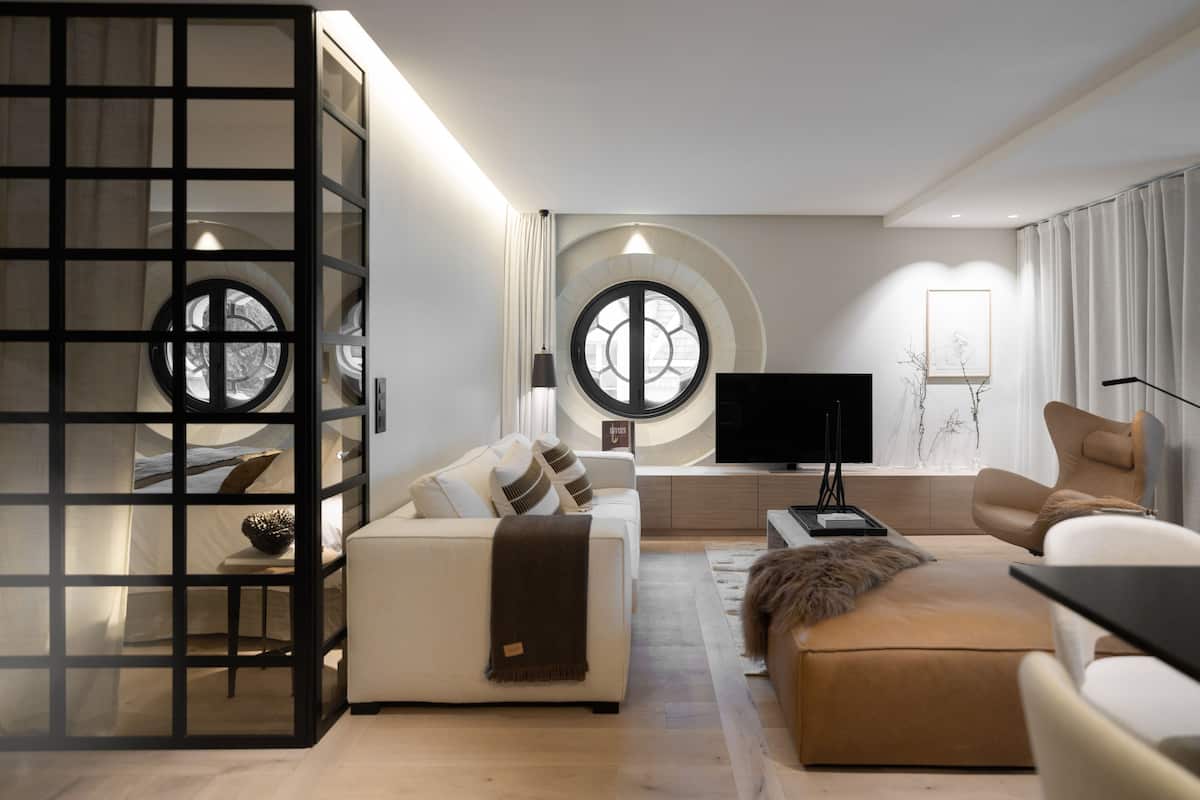 A modern living room with a sofa, ottoman, and a TV. There are two windows with circular designs, a black TV stand, and a decorative wall piece.