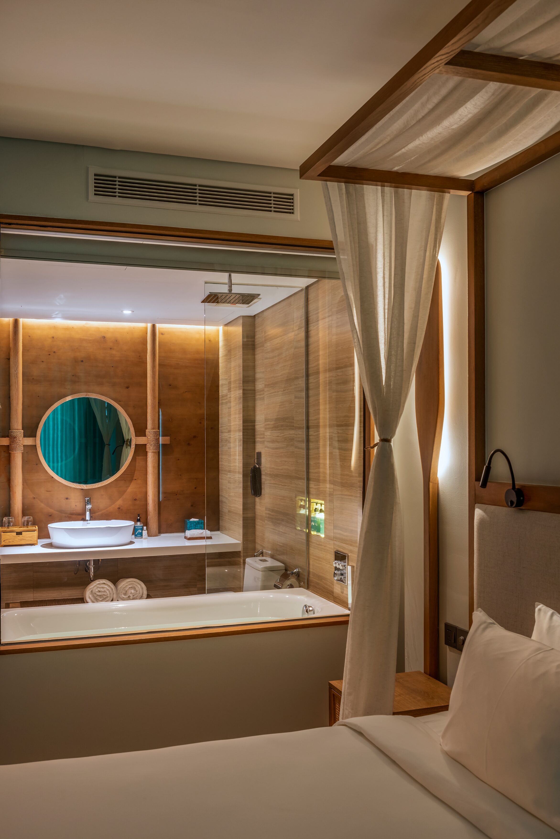 Habitación de hotel con una cama grande, cabecera de madera, espejo redondo, lavamanos y zona de ducha.