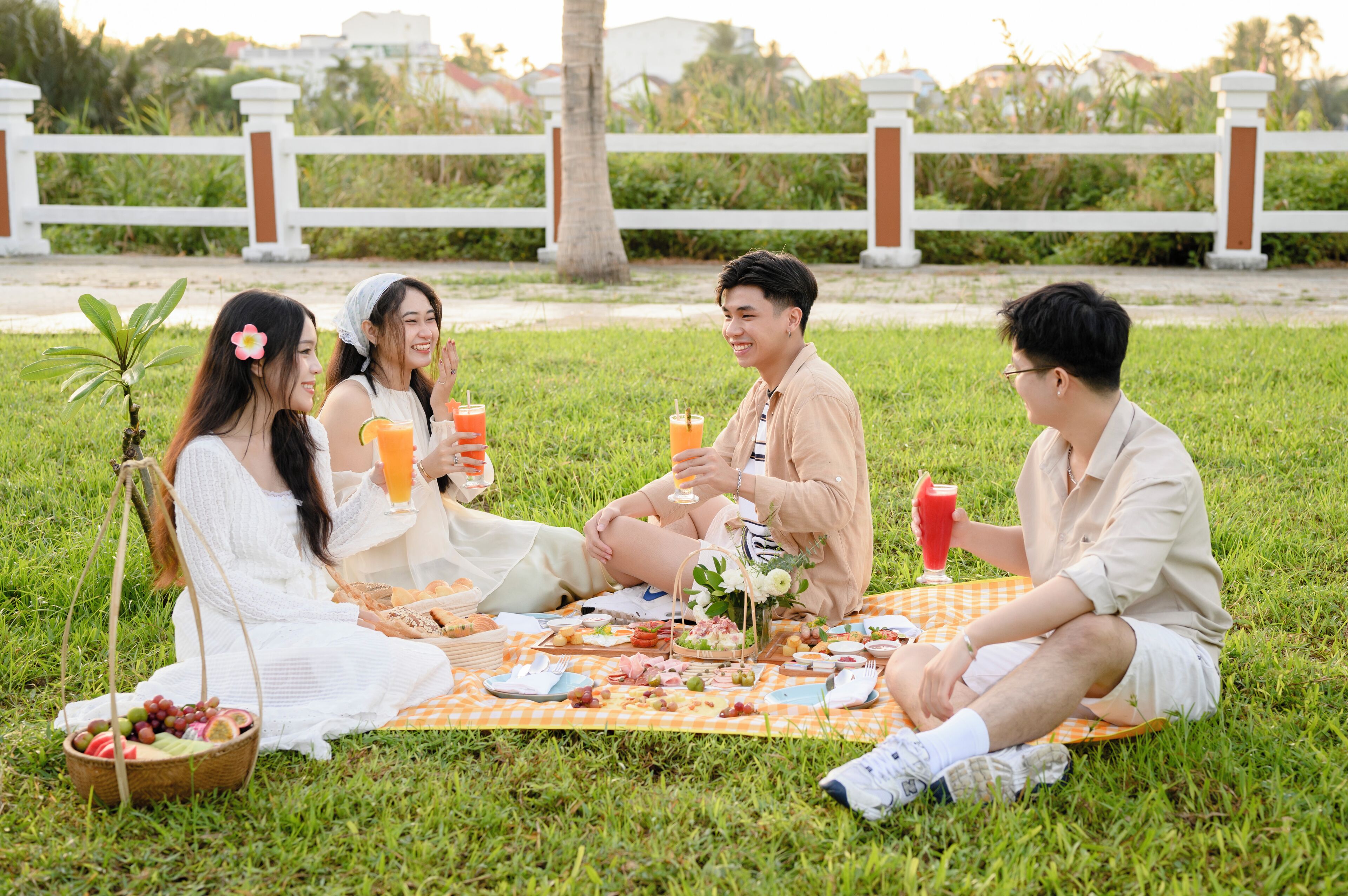A group of people having a picnic on a grassy area with a white fence and greenery in the background.