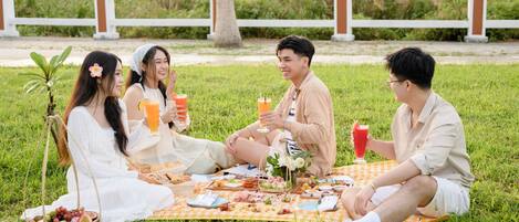 A group of people having a picnic on a grassy area with a white fence and greenery in the background.