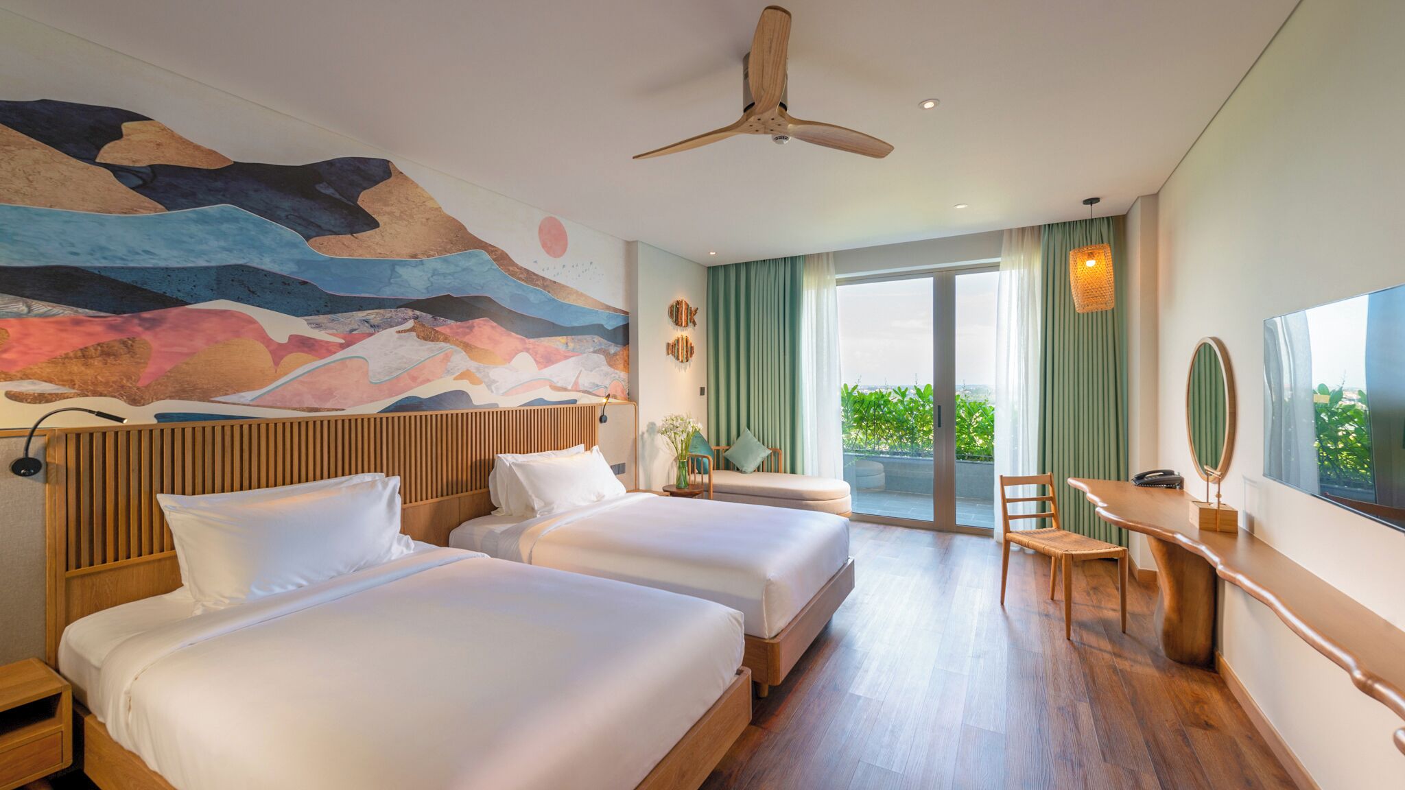 Une chambre d’hôtel avec deux lits, un bureau, une chaise, un ventilateur de plafond et une grande fenêtre donnant sur un paysage verdoyant.