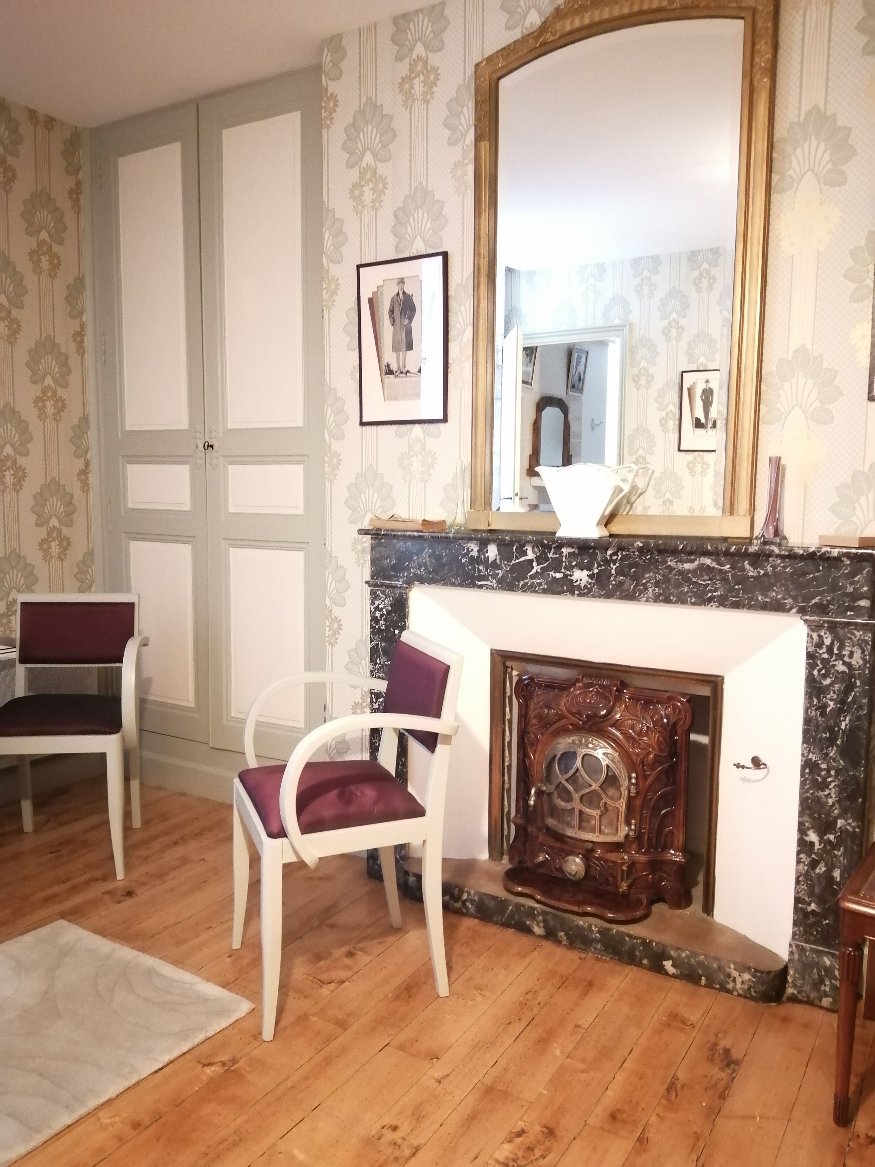 A room with a fireplace, two chairs, a mirror, and patterned wallpaper.