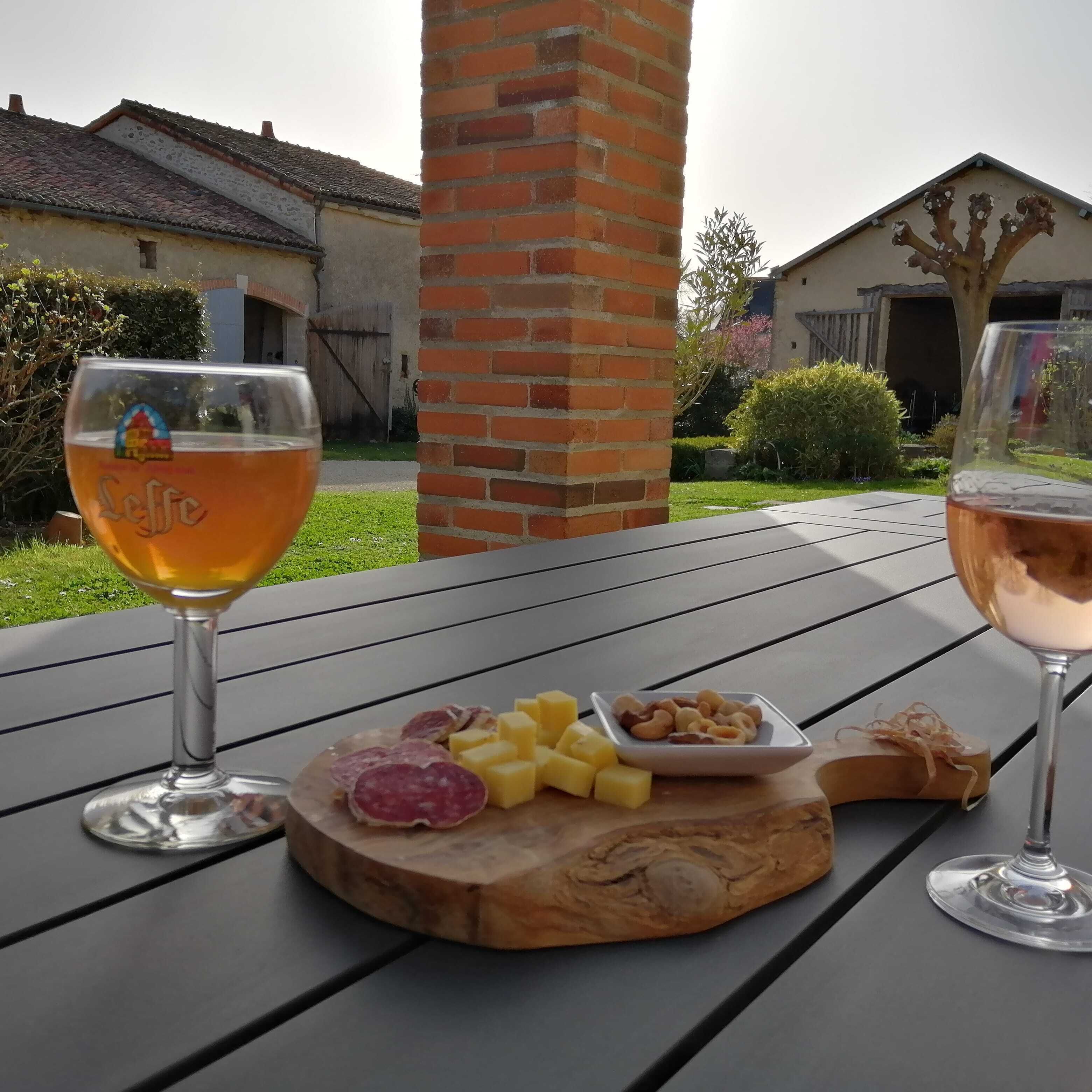 A table with two glasses of wine, a wooden board with cheese and cured meats, and a brick pillar.