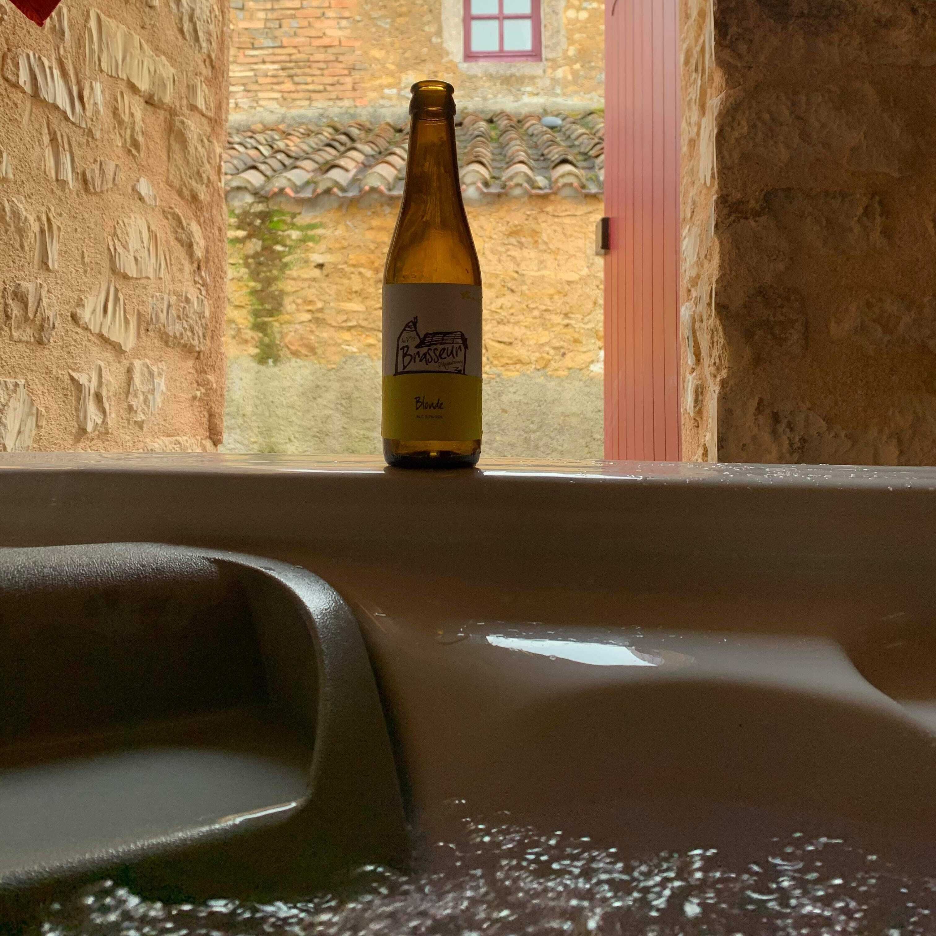 A bottle of Brasseur Blonde beer in a bathtub with a view of a stone building and a red door.