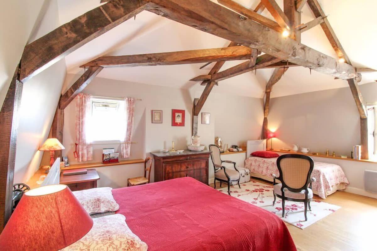 A bedroom with a sloped ceiling, wooden beams, a bed with red bedding, a desk with a chair, and a window with curtains.