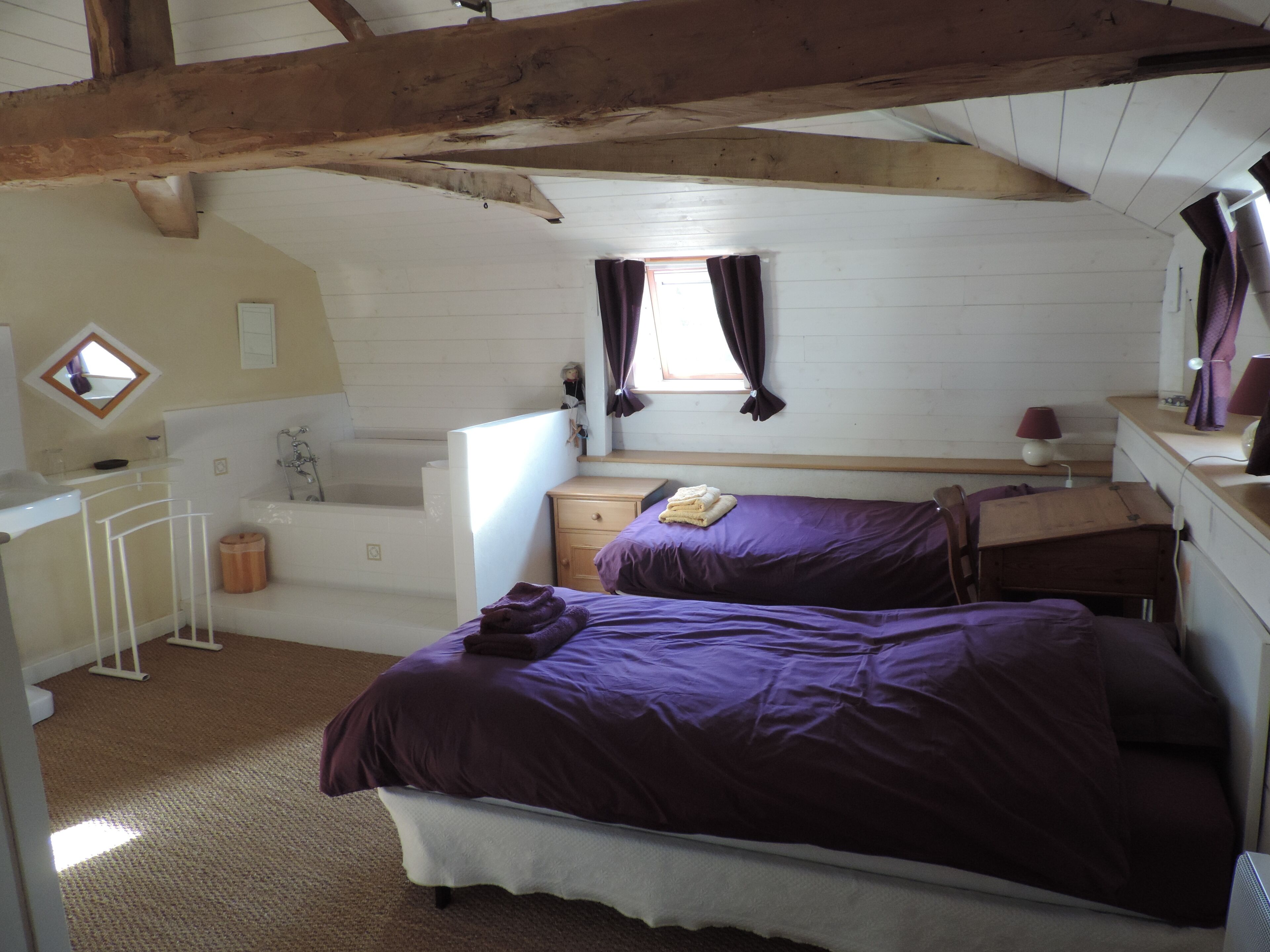 A bedroom with a large bed, a small desk, a chair, a window with curtains, a mirror, and a bathtub.