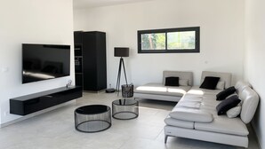 A modern living room with a flat-screen TV, a sofa set, a floor lamp, and a window with a view of trees. - LUXURIOUS ARCHITECT VILLA with OVERFLOW SWIMMING POOL and FOREST VIEW (Belin-Béliet)