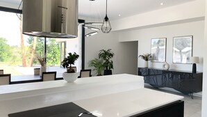 A modern kitchen with a central island, stainless steel range hood, pendant lighting, and a view of the outdoors. - LUXURIOUS ARCHITECT VILLA with OVERFLOW SWIMMING POOL and FOREST VIEW (Belin-Béliet)