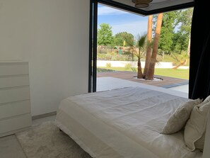 A bedroom with a large bed, a view of a garden with palm trees, and a modern design. - LUXURIOUS ARCHITECT-DESIGNED VILLA with an INFINITY POOL and a VIEW OF THE FOREST (Belin-Béliet)