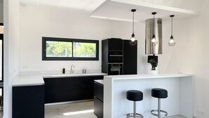 A modern kitchen with a white countertop, black cabinets, and a built-in oven. There are two bar stools and pendant lights above the counter. - LUXURIOUS ARCHITECT VILLA with OVERFLOW SWIMMING POOL and FOREST VIEW (Belin-Béliet)