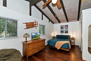 A bedroom with a wooden bed, a dresser, a desk, and a chair. There are windows with blinds, a ceiling fan, and framed pictures on the wall.