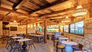 A rustic interior with wooden beams, stone walls, and a bar area with a view. Tables and chairs are arranged for dining.