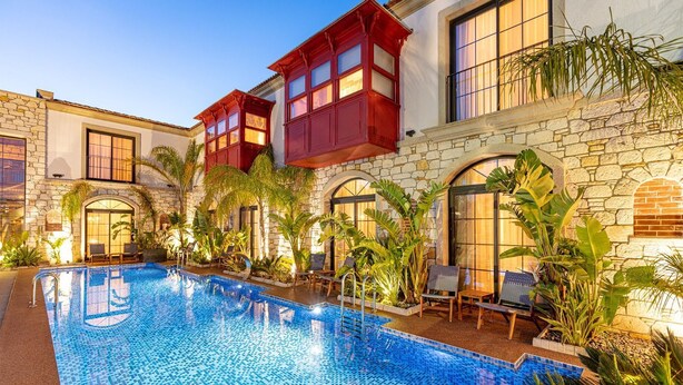 A poolside area with a clear blue pool, stone walls, and red wooden balcony railings.