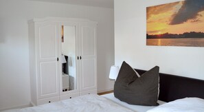 Un dormitorio con una cama, un armario blanco, una mesita de noche, una lámpara y un cuadro de una puesta de sol sobre el agua.