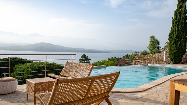 A poolside area with wicker chairs, a stone wall, and a view of the sea and mountains. - Exceptional Corsican villa in the Gulf of Porto-Vecchio with its feet in the water (Porto-Vecchio)