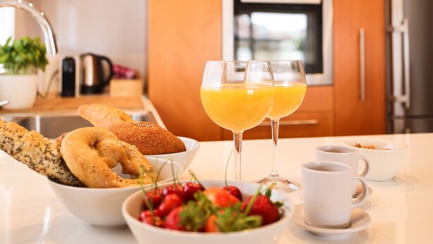 A breakfast setting with bagels, a bowl of strawberries, two glasses of orange juice, and a coffee cup on a kitchen counter.
