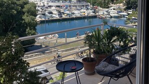 A balcony with wicker chairs, a small table, and potted plants. It overlooks a marina with boats and a clear blue sky.