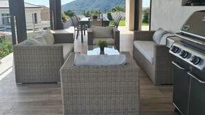 A modern outdoor living area with wicker furniture, a barbecue grill, and a view of a mountain.