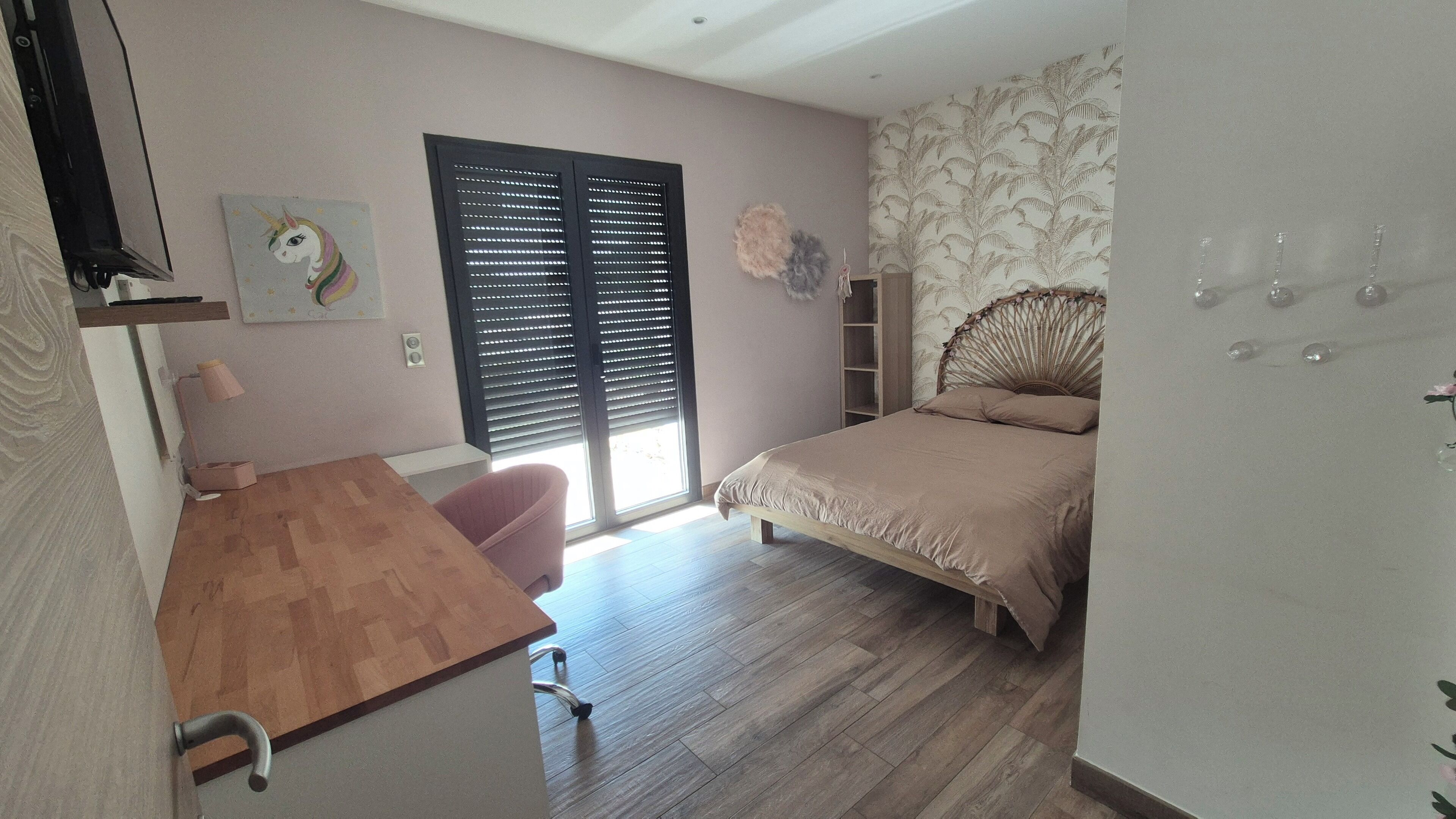 Une chambre avec un bureau en bois, un lit doté d’une tête de lit en osier, une fenêtre avec des stores et un mur recouvert d’un papier peint à motifs floraux.
