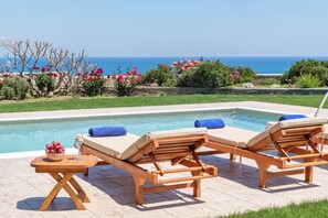 A poolside area with two wooden lounge chairs, a small round table with a bowl of fruit, and a view of the sea.