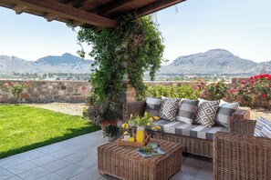 Ein Korbesofa mit Kissen, ein Glastisch mit einer Flasche und Blumen sowie ein Blick auf Berge.