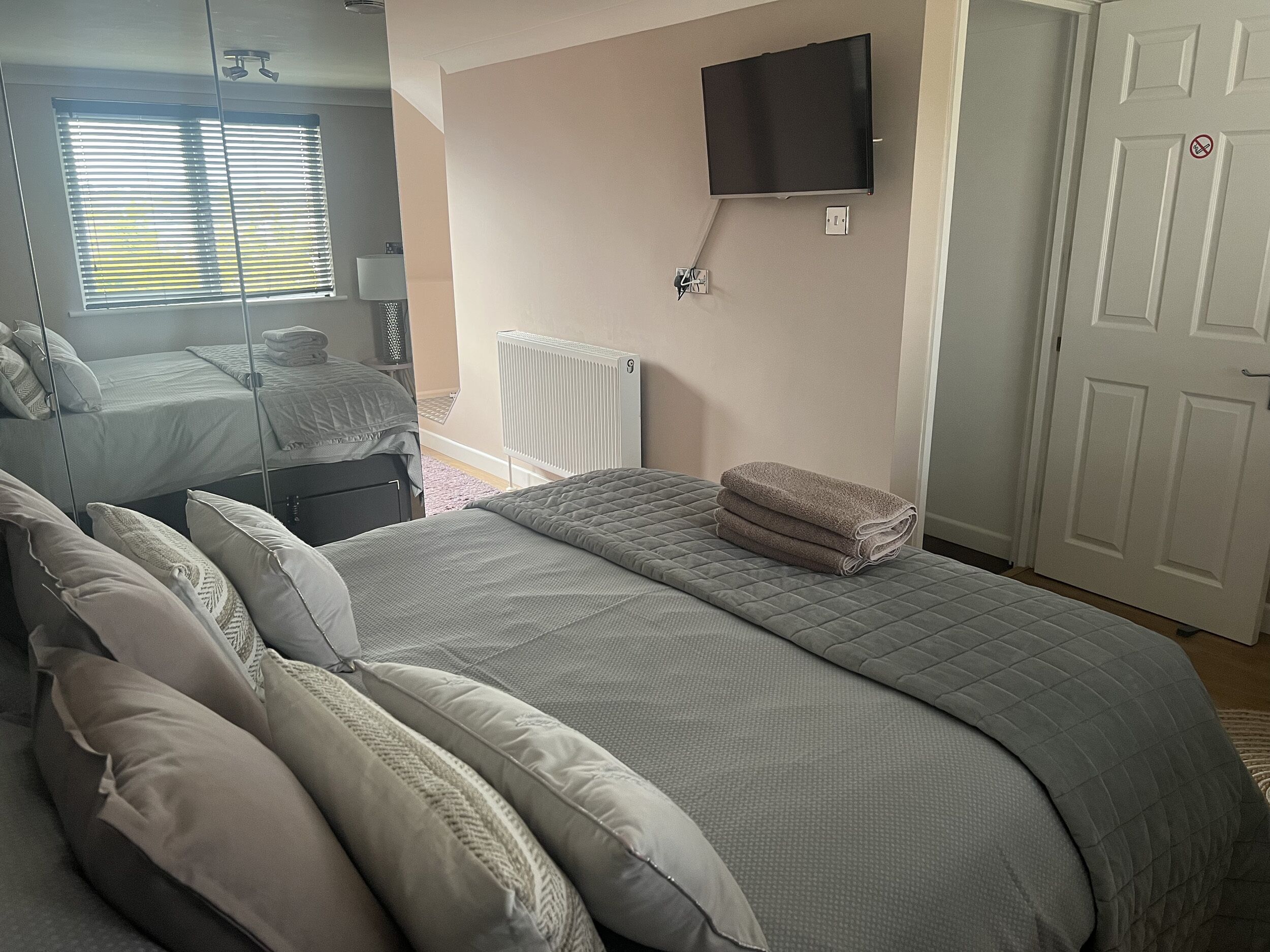 A bedroom with a bed, a TV mounted on the wall, a radiator, and a door with a no-smoking sign.