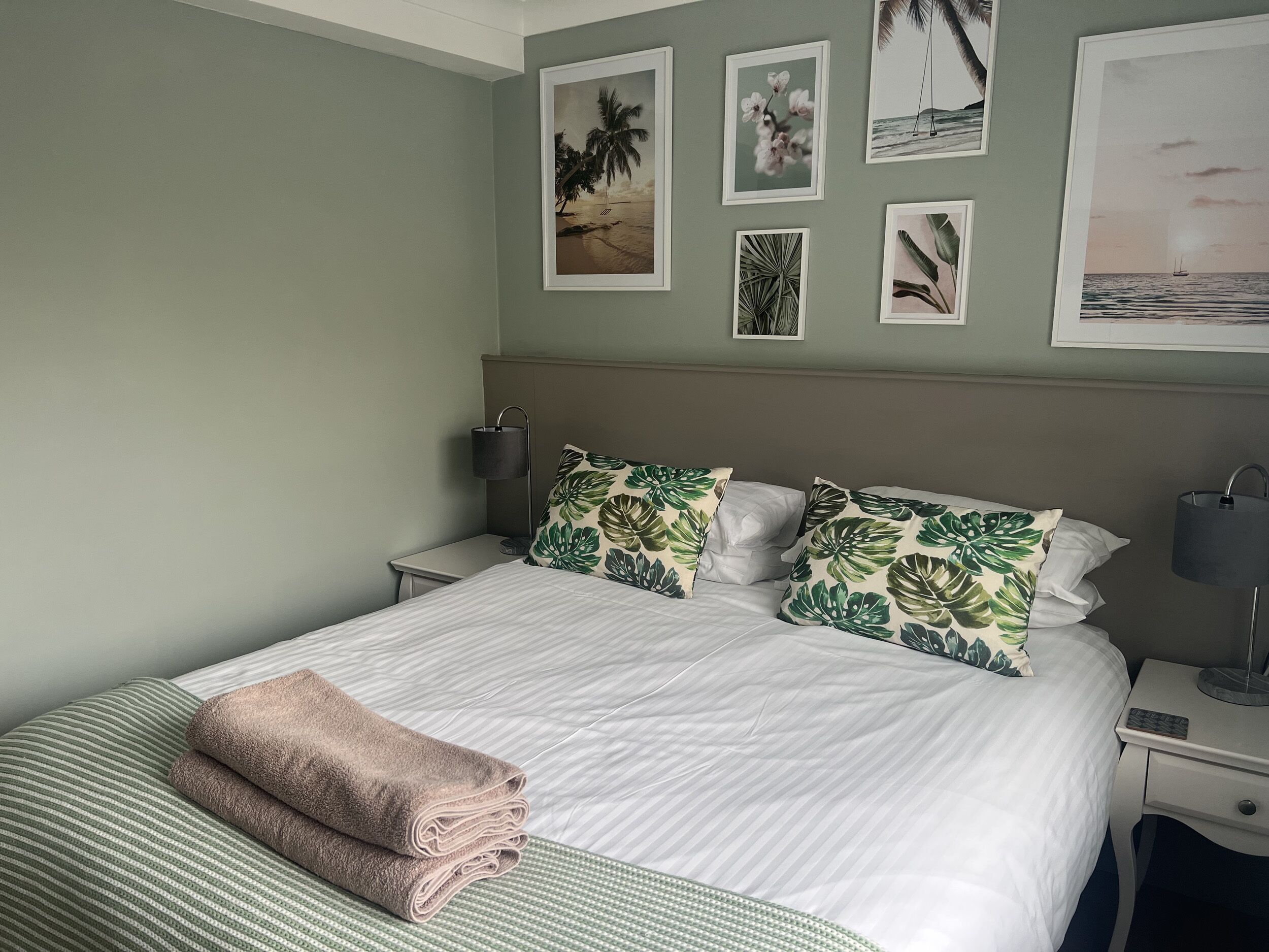 A neatly made bed with a striped pillow and two folded towels, a nightstand with a lamp, and a wall with framed tropical-themed artwork.