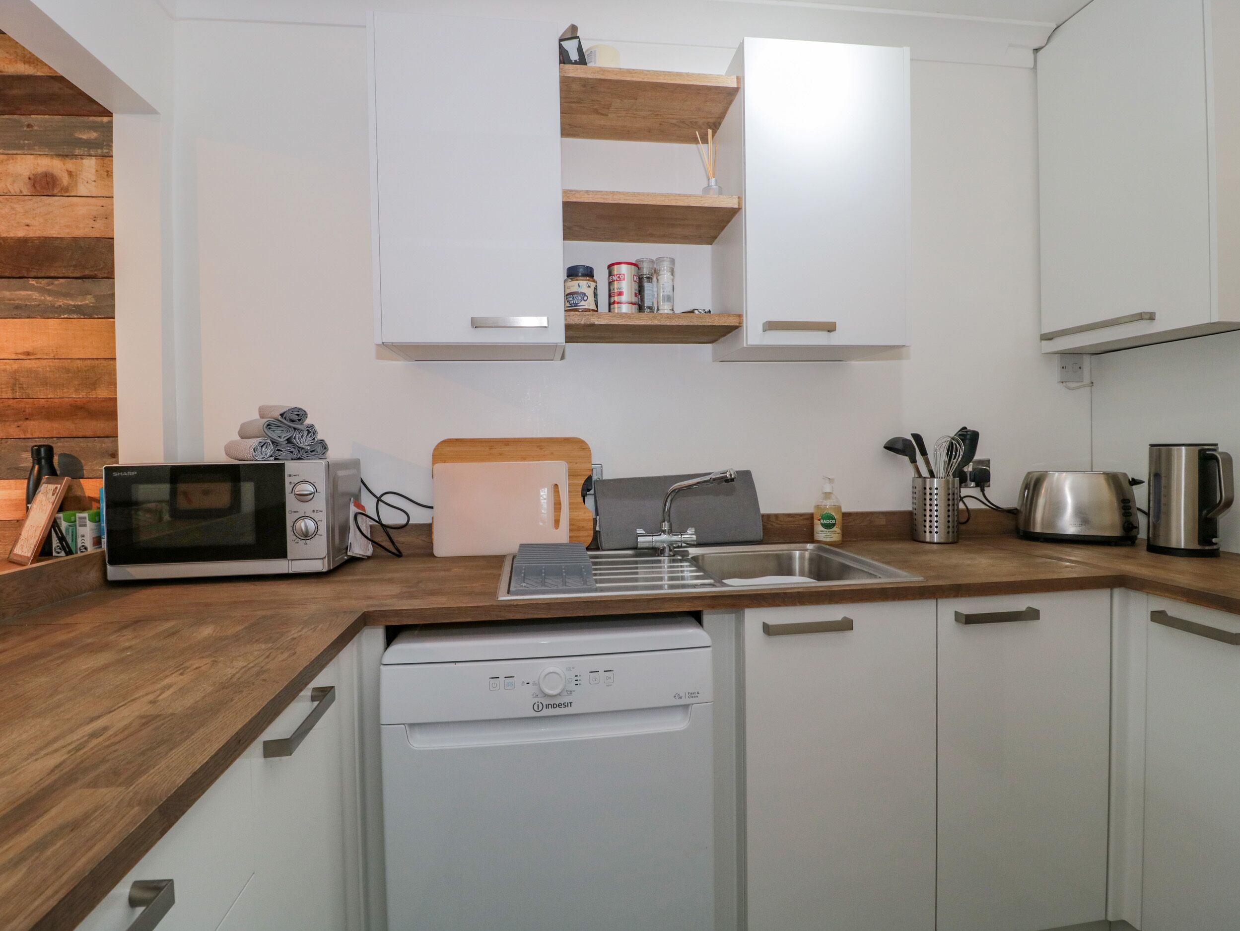 Una cocina moderna con armarios blancos, encimera de madera, lavavajillas integrado, microondas, tostadora y un lavabo.