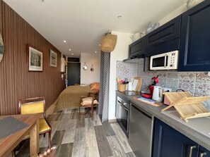 A compact kitchen with a microwave, toaster, and wooden drying rack. A bed with a brown blanket and pillows is visible in the background. - Renovated studio with AC and wifi at Aiguillon port for 2 (Arcachon)