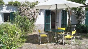 An outdoor patio with a table and chairs under a white umbrella, surrounded by greenery and a white building with green shutters. - The Sea - White House (Camporosso)