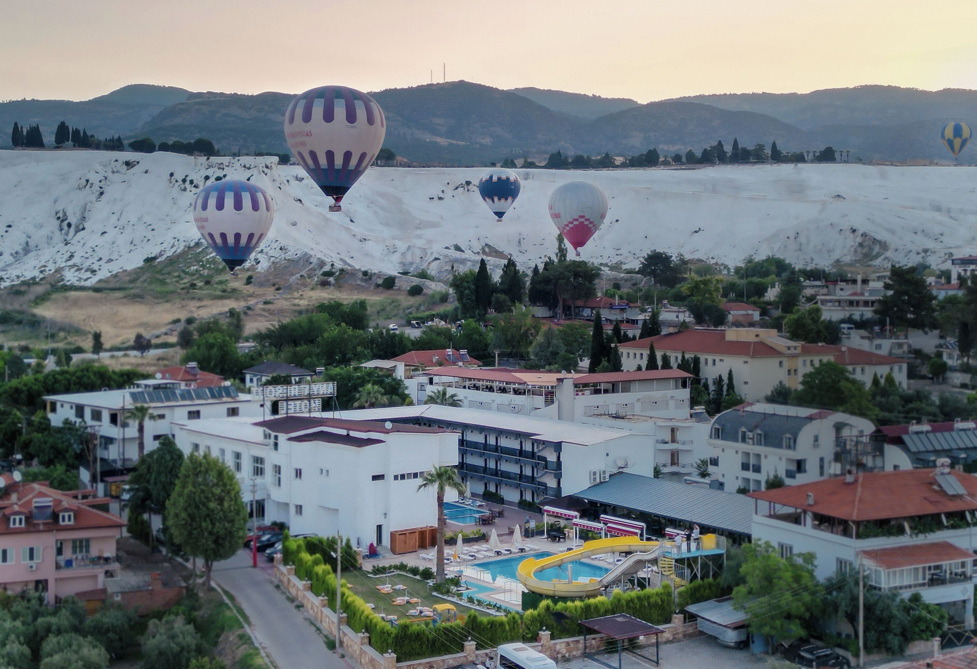 Un resort con varias edificaciones, una piscina y globos aerostáticos al fondo.