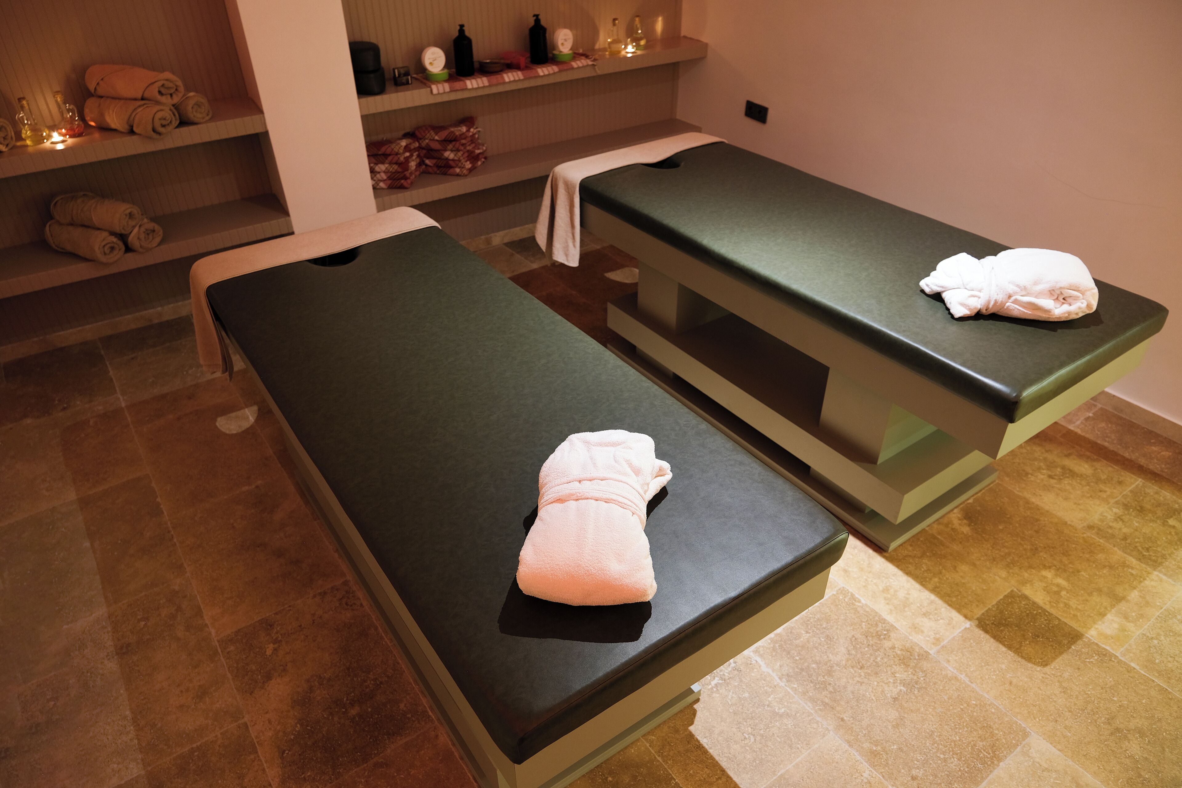 Duas mesas de massagem num espaço com chão em mosaico, uma estante com toalhas e produtos de higiene, e uma prateleira na parede com frascos.