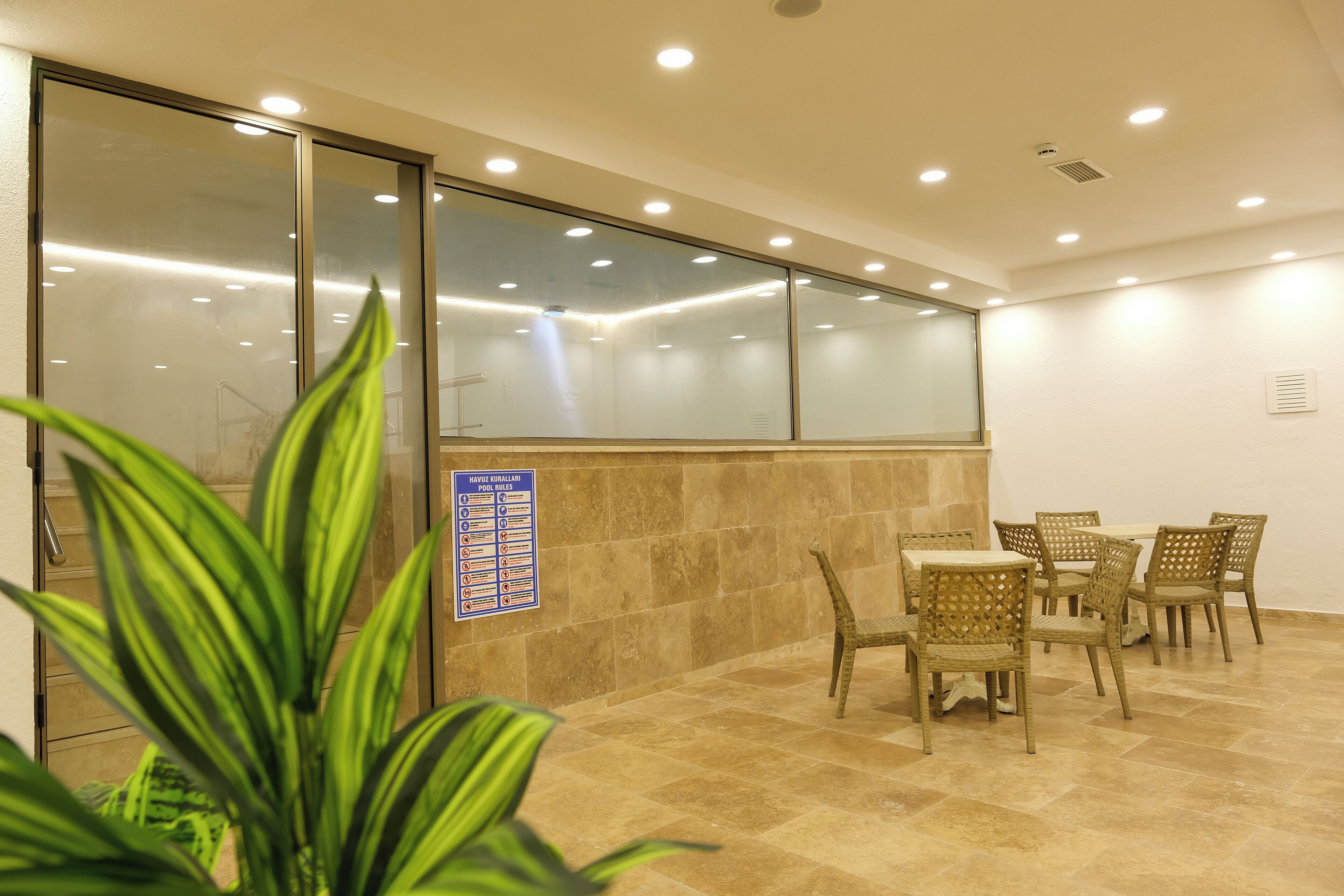 Habitación de hotel con separador de vidrio, un cartel con las normas de la piscina, zona de comedor con sillas de mimbre y suelo de baldosas.