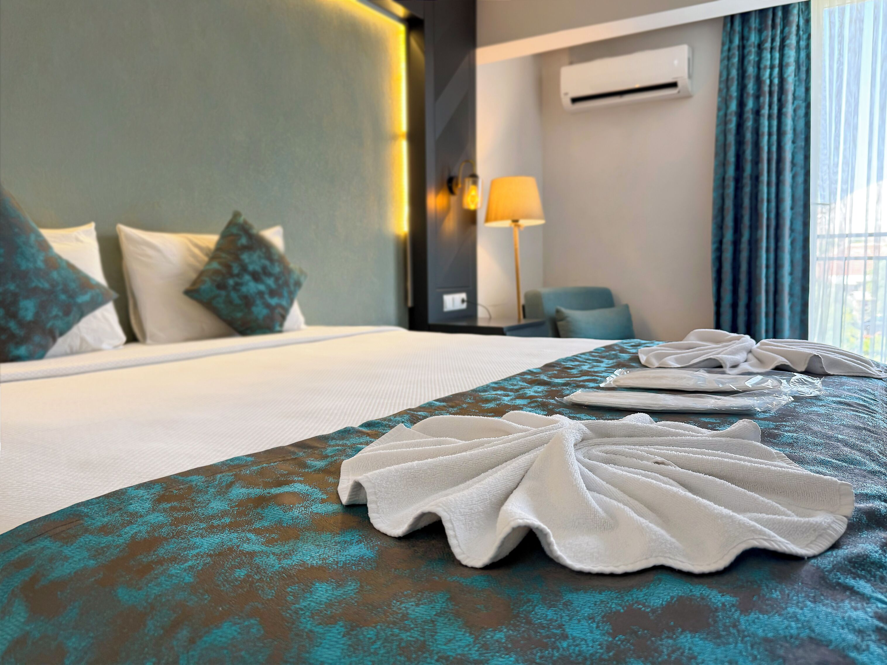 A hotel room with a neatly made bed, a bedside table with a lamp, and a view of the city through the window.
