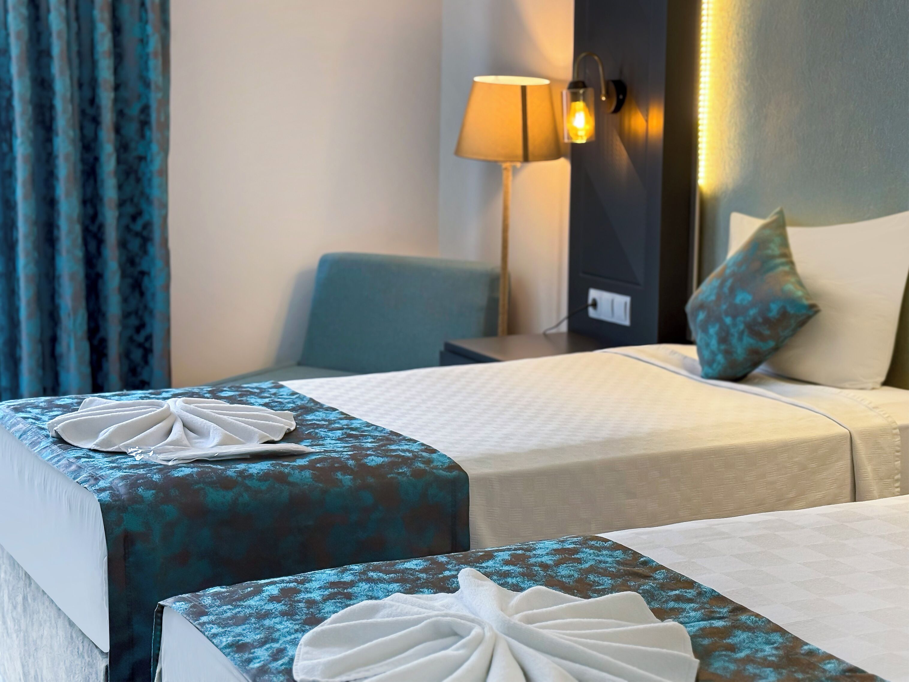 A hotel room with two beds, each with a patterned bedspread and a white towel. There is a lamp on the wall and a chair in the corner.