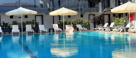 Une piscine entourée de chaises longues blanches et de parasols, le tout bordé d’un bâtiment moderne doté de balcons et d’un escalier.