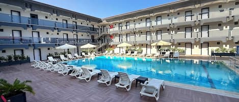 Zona de piscina de hotel con múltiples sillas de descanso y sombrillas, rodeada por un edificio de varios pisos con balcones.